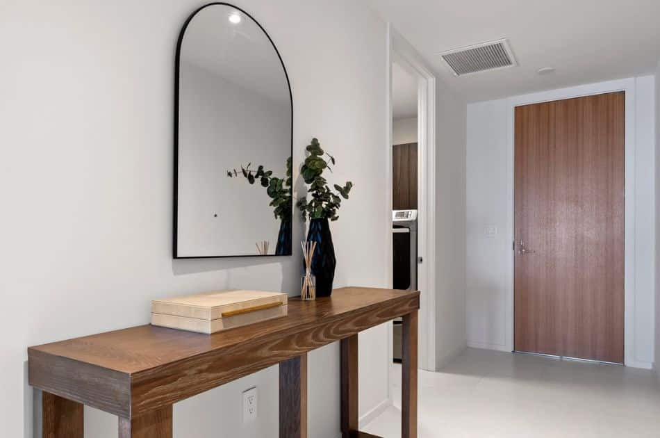 A modern entryway featuring a wooden console table topped with a decorative box, a vase with greenery, and a reed diffuser. A large, arched mirror is mounted on the wall above the table, reflecting natural light from the minimalist space.