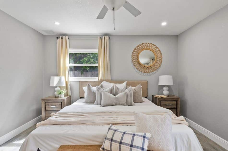 A cozy bedroom designed with soft neutral tones, featuring a neatly made bed with layered pillows and a throw blanket. Two matching bedside tables with lamps frame the bed, and a circular mirror decorates the wall above the headboard.