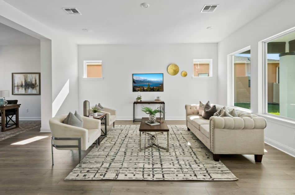 A bright and spacious living room with large windows, showcasing neutral-toned furniture including a tufted sofa, two accent chairs, and a patterned rug. The space is styled with modern decor and a wall-mounted TV as the focal point. | small florida condo decorating ideas