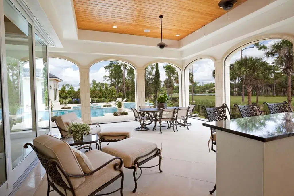 A luxurious covered patio with arched openings overlooking a scenic backyard and pool, furnished with elegant wrought iron chairs, a lounge chair, and an outdoor kitchen area with a granite countertop.