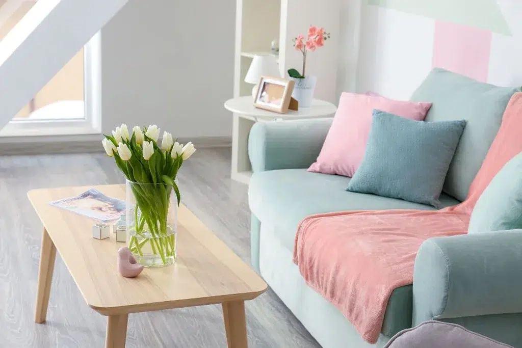 A charming living room with a pastel aesthetic, featuring a mint-green sofa accented with pink and blue throw pillows, a light wood coffee table with a vase of white tulips, and a bright, airy atmosphere.