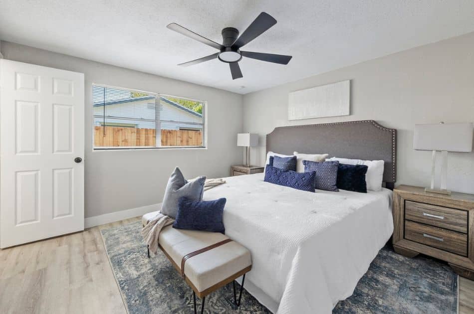 A cozy bedroom featuring a bed with a gray studded headboard, crisp white bedding, and navy accent pillows. The room includes matching wooden nightstands with modern lamps and a stylish bench at the foot of the bed, complemented by light wood floors and a ceiling fan.