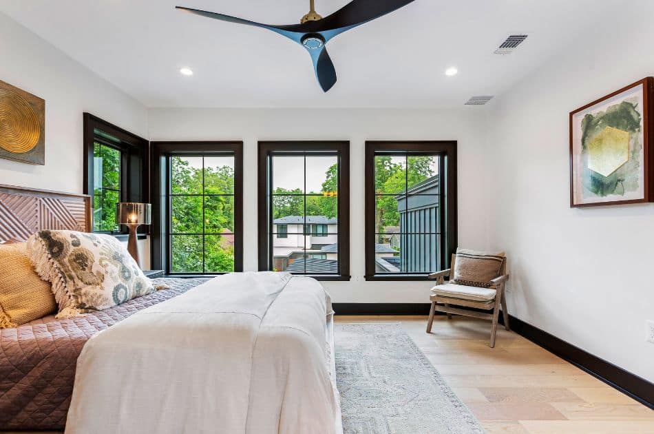 A bright and airy bedroom with large black-trimmed windows offering outdoor views. The room highlights a bed with a patterned wooden headboard, soft textured bedding, and decorative pillows. A single chair and artwork add character, with a sleek ceiling fan overhead.