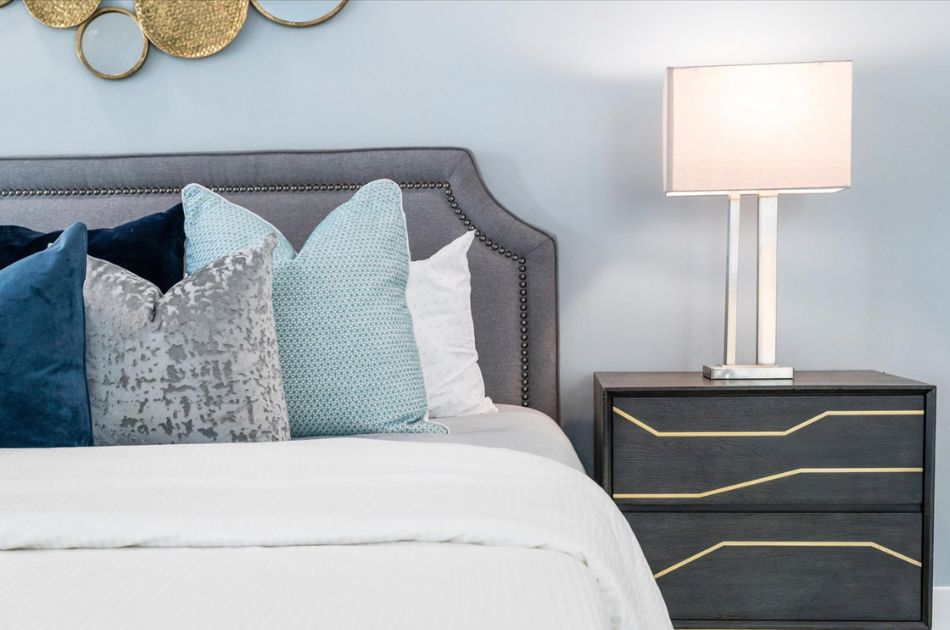 Close-up of a bed with blue and gray throw pillows, a dark nightstand, and a modern table lamp.